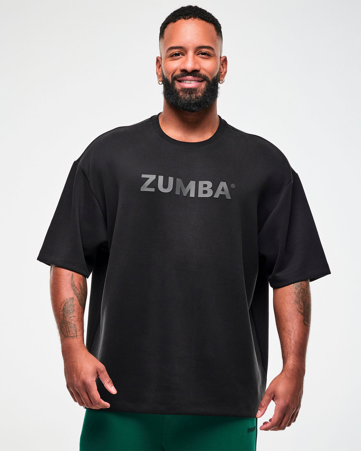Male model in a black oversized drop shoulder crew neck top with large dark 'ZUMBA' text across the chest, paired with teal green pants.