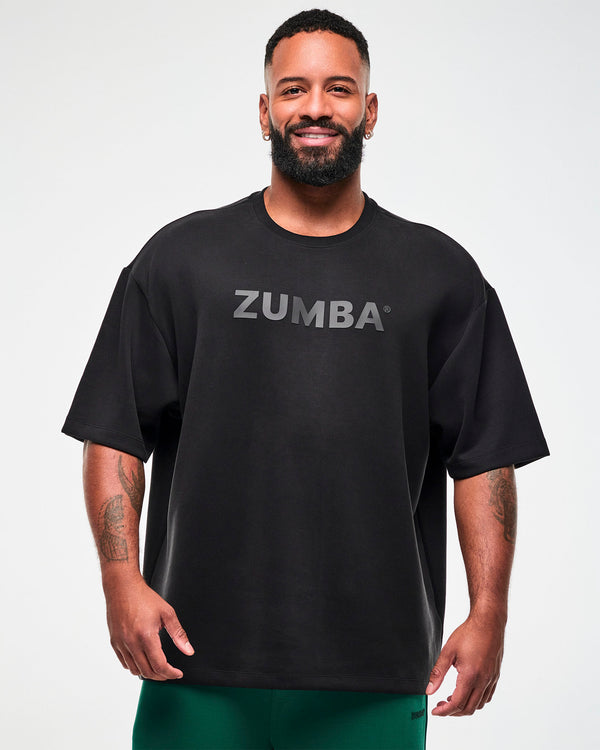 Male model in a black oversized drop shoulder crew neck top with large dark 'ZUMBA' text across the chest, paired with teal green pants.