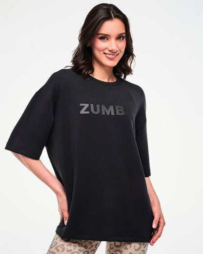 Female model in a black oversized drop shoulder crew neck top with large dark 'ZUMBA' text across the chest, paired with beige leopard print biker shorts.