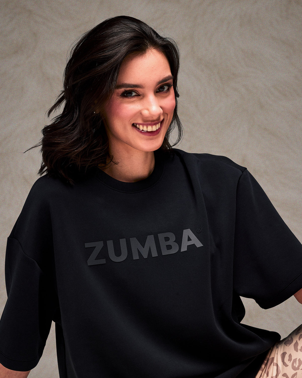 Close-up of a smiling female model in the black oversized top with large dark 'ZUMBA' text across the chest. Plain taupe backdrop.