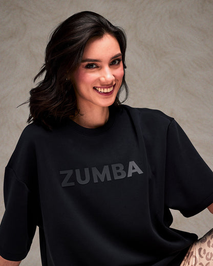 Close-up of a smiling female model in the black oversized top with large dark 'ZUMBA' text across the chest. Plain taupe backdrop.
