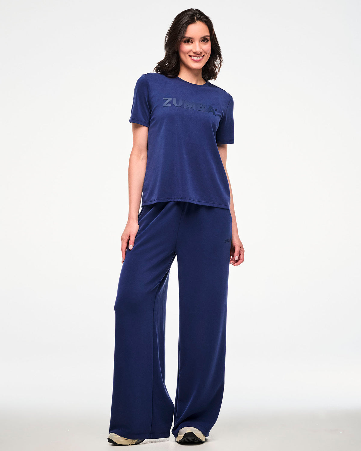 Full view of a female model in a navy crew neck top with large tonal 'ZUMBA' text across the chest, paired with matching navy high-waisted straight-leg pants and beige shoes.