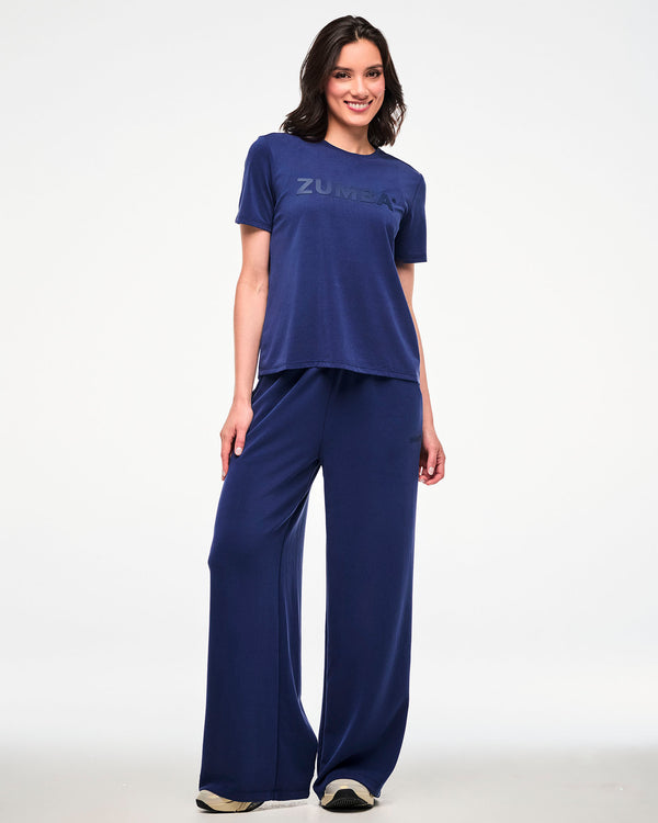 Full view of a female model in a navy crew neck top with large tonal 'ZUMBA' text across the chest, paired with matching navy high-waisted straight-leg pants and beige shoes.
