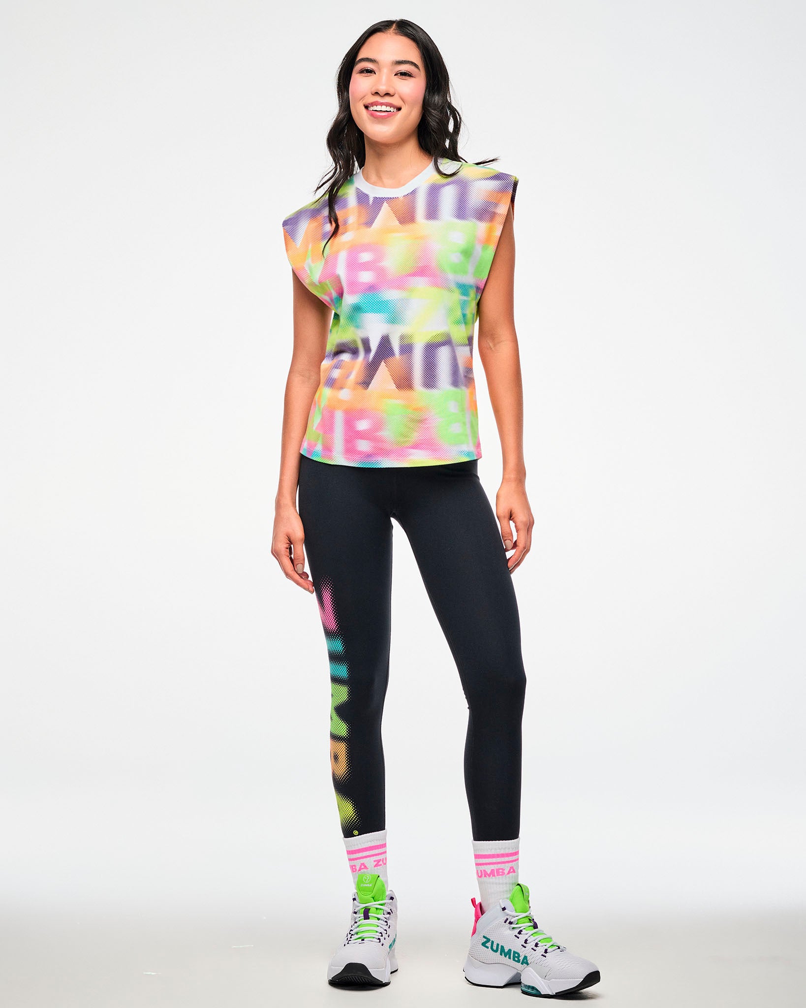 Female model wearing a multicolor halftone print extended shoulder tank, paired with black ZUMBA leggings, pink striped socks, and white sneakers.
