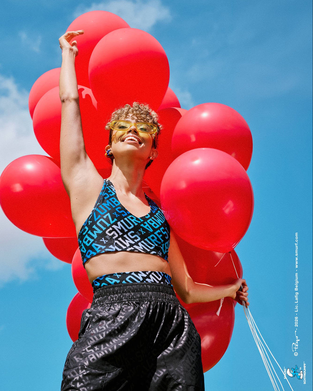 Female model holding a cluster of red balloons, wearing a reversible V-neck racerback bra print side out, with diagonal blue and white 'ZUMBA' X 'THE SMURFS' varsity lettering on black, paired with black tonal print pants against a blue sky.
