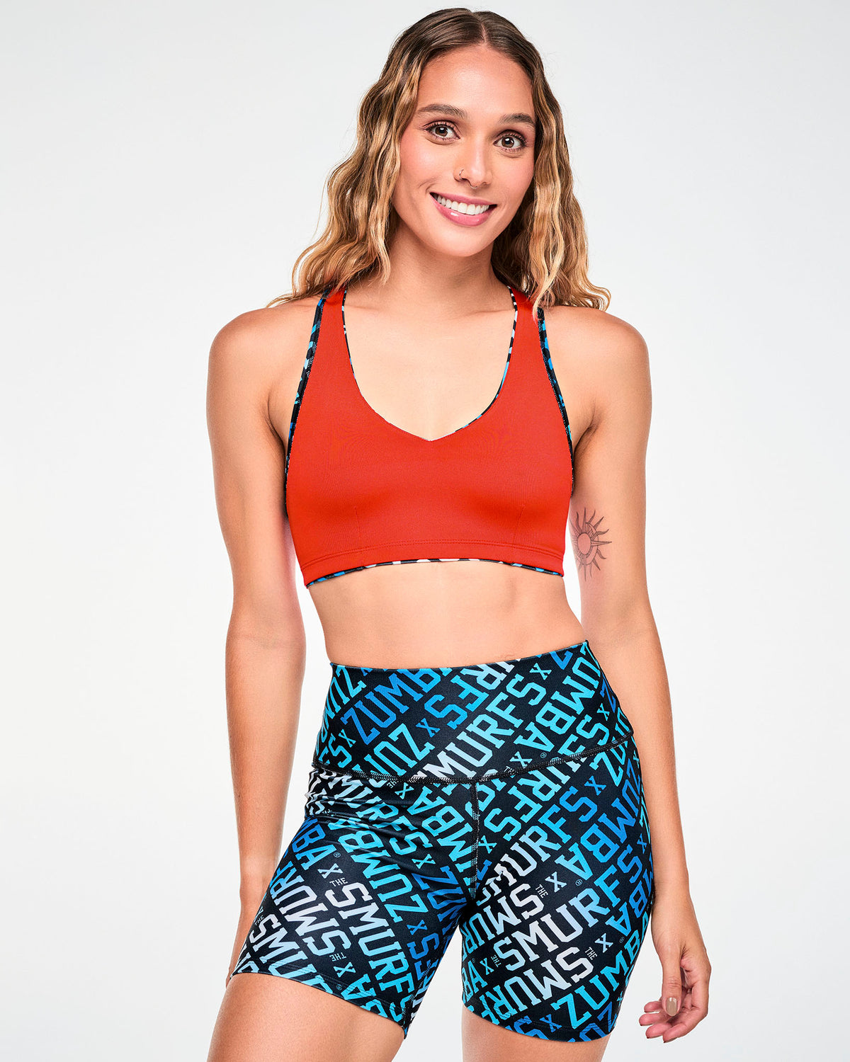 Female model in a reversible V-neck racerback bra worn red side out, with the blue, white, and black 'ZUMBA' X 'THE SMURFS' print side showing at the edges. Paired with matching print biker shorts.