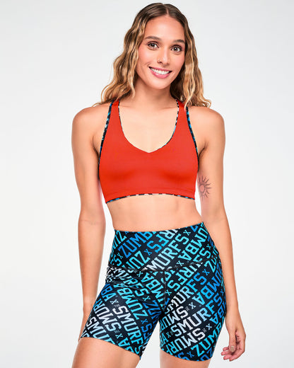 Female model in a reversible V-neck racerback bra worn red side out, with the blue, white, and black 'ZUMBA' X 'THE SMURFS' print side showing at the edges. Paired with matching print biker shorts.