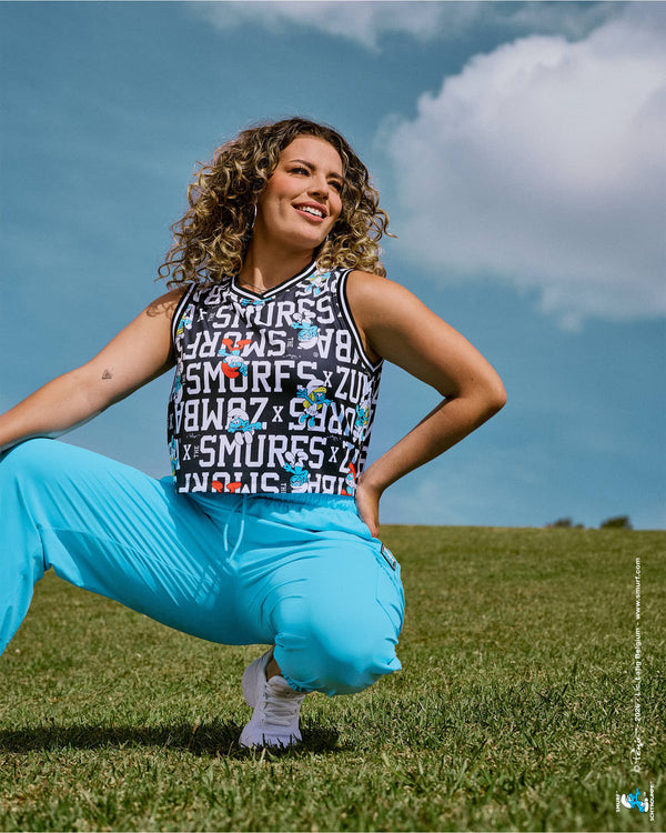 Female model crouching on a grassy field in the black and white Smurfs character print crop tank with striped trim, paired with aqua blue cargo pants under a cloudy sky.