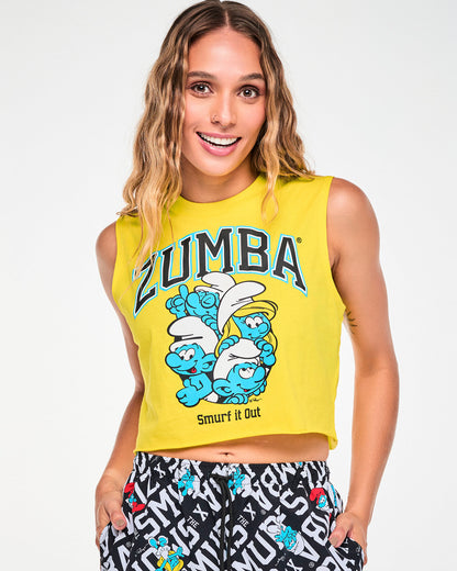 Female model in a yellow sleeveless crew neck crop tank with a raw-edge hem, featuring arched 'ZUMBA' text above a group of Smurfs, including Brainy and Smurfette, and 'Smurf it Out' text. Paired with Smurfs print cargo pants.
