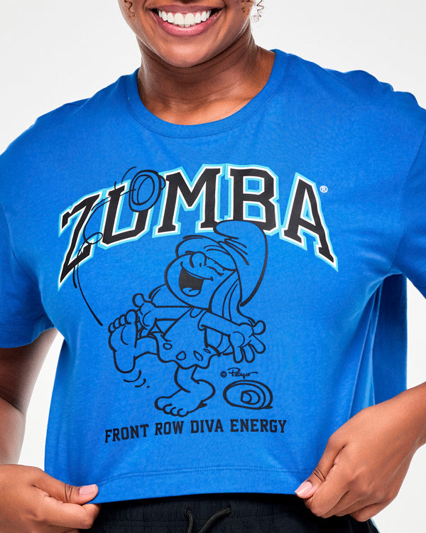 Close-up of the blue crop top's graphic: black varsity 'ZUMBA' text outlined in light blue above a black line drawing of Smurfette laughing, kicking off her shoes and dancing barefoot, with 'FRONT ROW DIVA ENERGY' below.