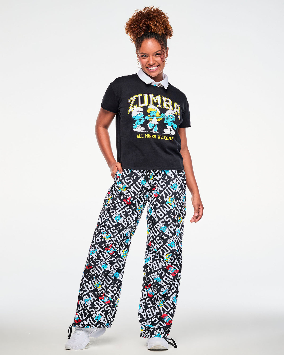 Female model with one hand in her pocket, wearing the black Smurfs graphic top with 'ZUMBA,' three dancing Smurfs, and 'ALL MOVES WELCOME' over a white-collared mesh polo, paired with black and white Smurfs print cargo pants.