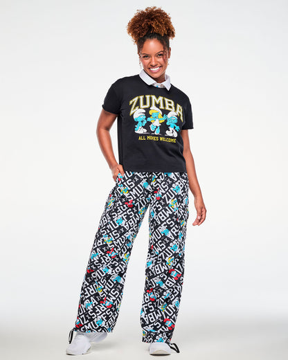 Female model with one hand in her pocket, wearing the black Smurfs graphic top with 'ZUMBA,' three dancing Smurfs, and 'ALL MOVES WELCOME' over a white-collared mesh polo, paired with black and white Smurfs print cargo pants.