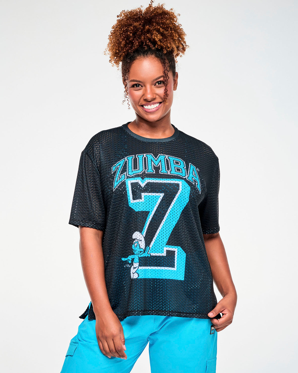 Female model in a black open-hole mesh boxy tee with side slits, featuring arched light blue 'ZUMBA' text over a large light blue varsity 'Z' outlined in white, with a Smurf leaning on the bottom of the letter. Paired with aqua blue cargo pants.