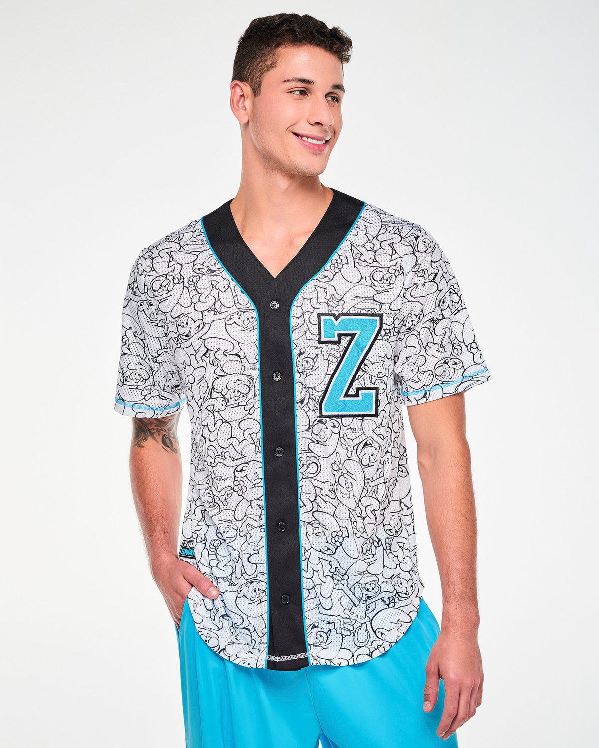 Male model in a white mesh baseball jersey printed all over with black line-drawn Smurfs, with a black V-neck placket and black buttons edged in light blue piping. A light blue varsity 'Z' outlined in black and white sits on the left chest, with a black 'ZUMBA' X 'THE SMURFS' patch near the right hem. Paired with aqua blue cargo pants.