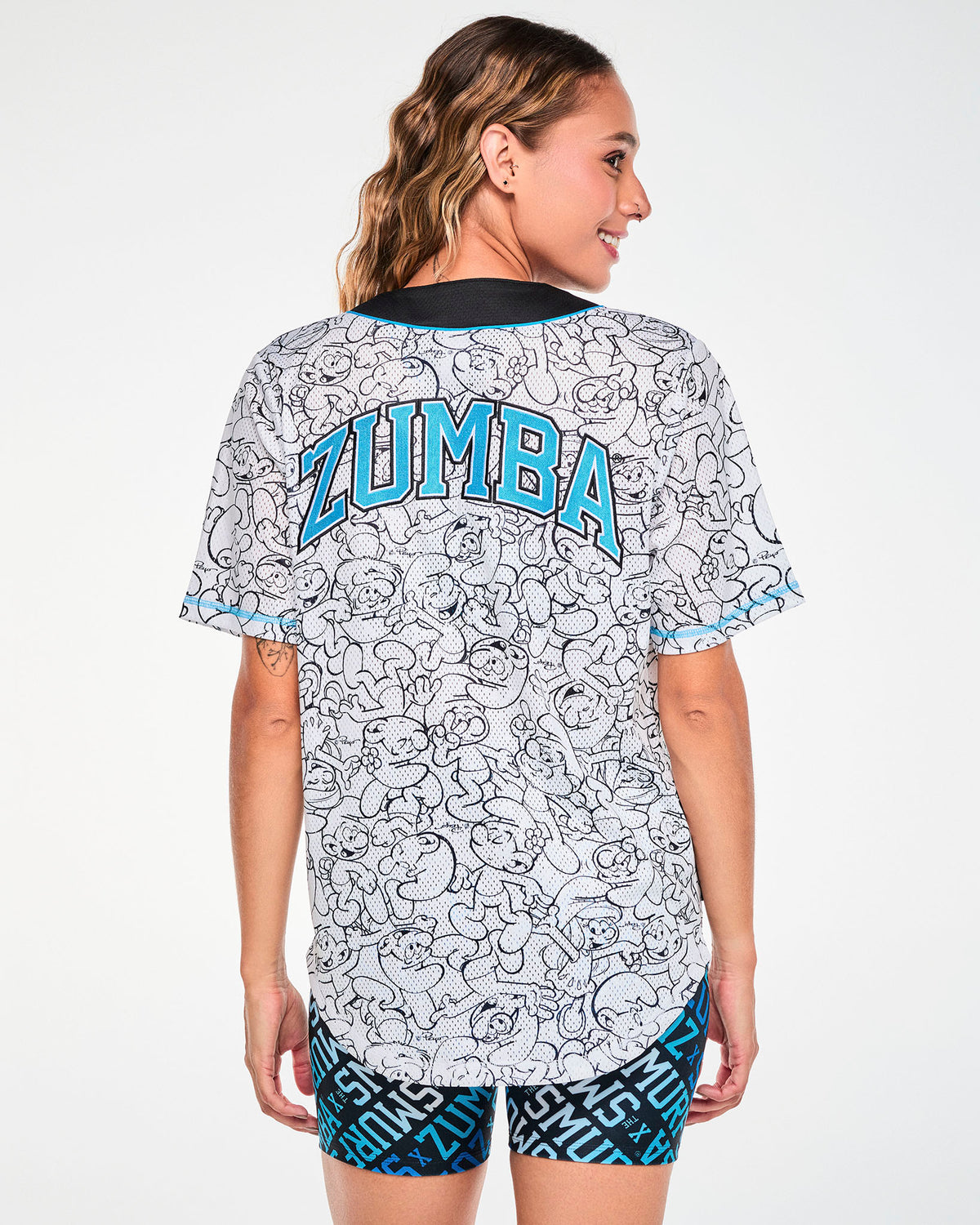 Shown from the back, a female model in the white Smurfs line-art baseball jersey, with the embroidered arched 'ZUMBA' text in aqua across the shoulders and the Smurfs line art continuing down to the rounded hem.