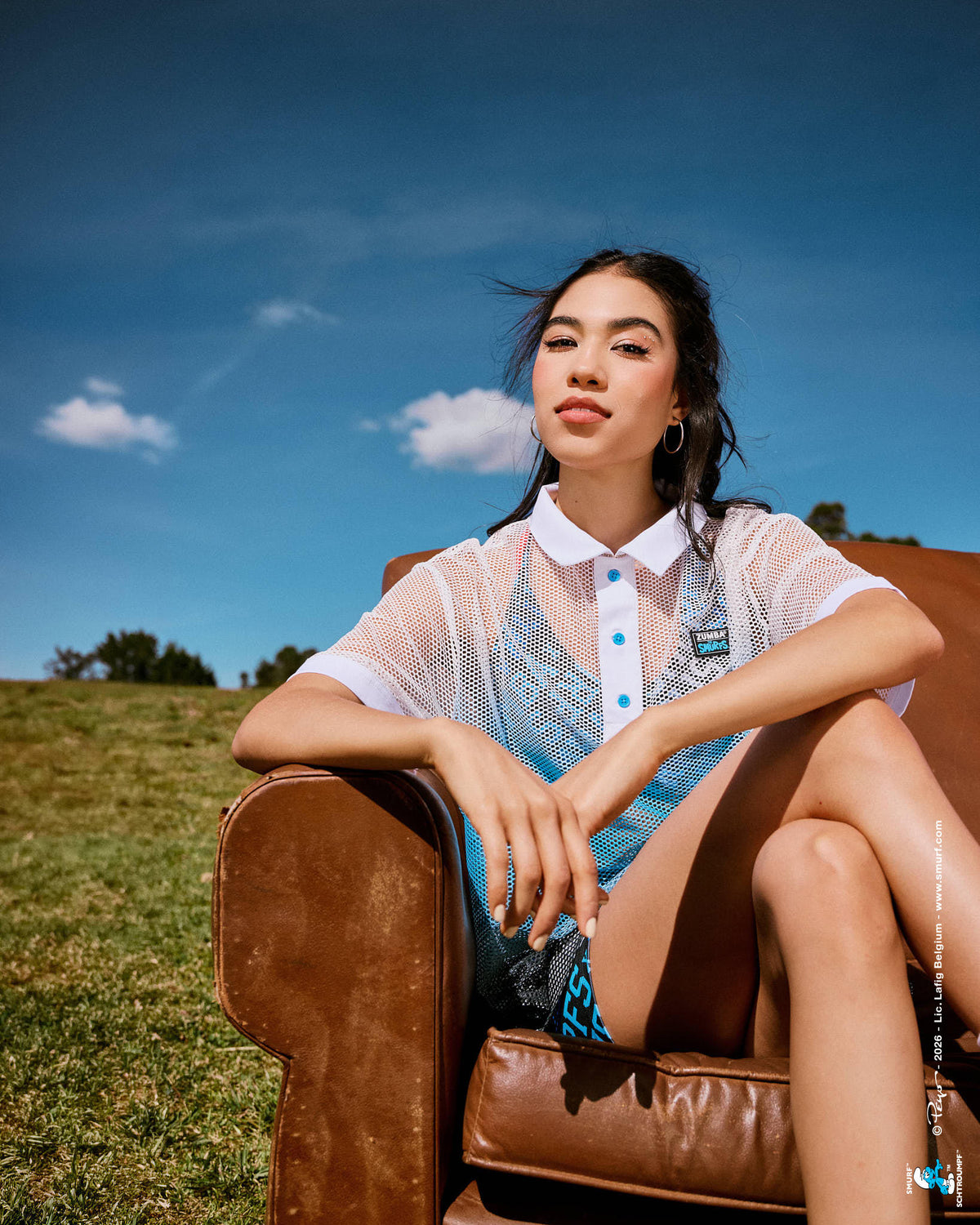 Female model lounging in a worn brown leather armchair on a grassy hill, wearing the white-to-blue mesh polo over a logo print bra, with a blue cloudy sky overhead.