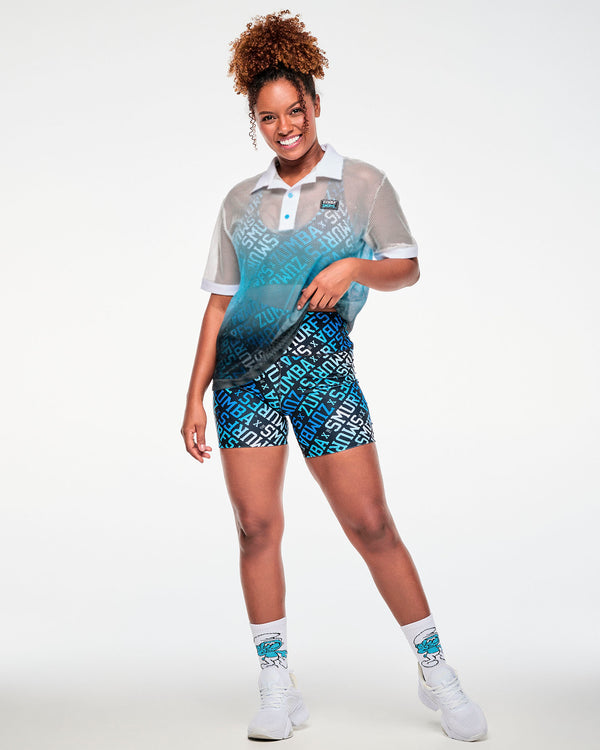 Female model pulling up the hem of the white-to-blue mesh polo, worn over a blue logo print bra with matching biker shorts, Smurf socks and white shoes.