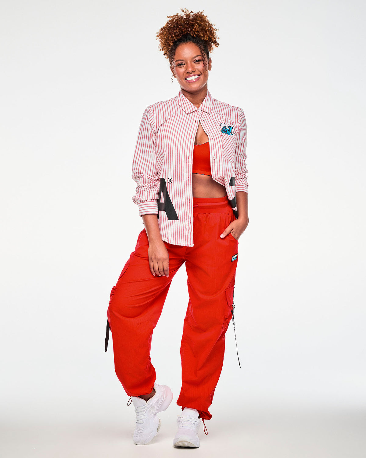 Female model in the red and white striped button-up worn open over a red bra, showing the embroidered Smurf on the chest pocket, paired with red cargo pants with black ‘ZUMBA’ straps and white shoes.