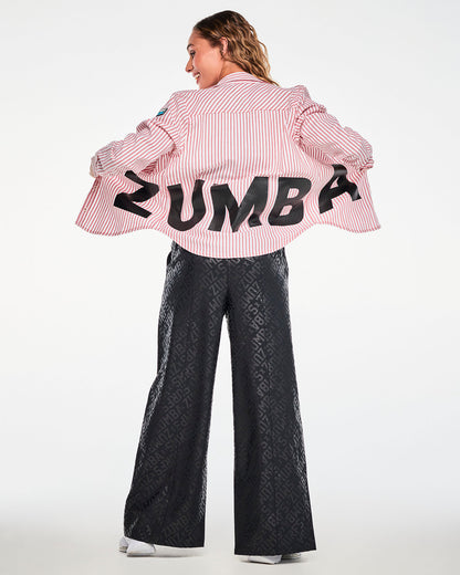 Shown from the back, a female model holding open the red and white striped button-up to reveal large black 'ZUMBA' letters across the lower back, paired with black tonal logo print wide-leg pants.
