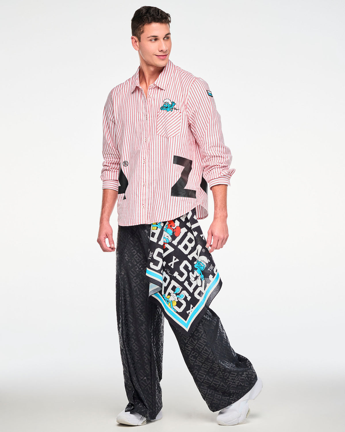 Male model in the red and white striped button-up with the embroidered Smurf on the chest pocket and black 'ZUMBA' letters at the hem, paired with black tonal logo print wide-leg pants and a Smurfs bandana hanging at the hip.