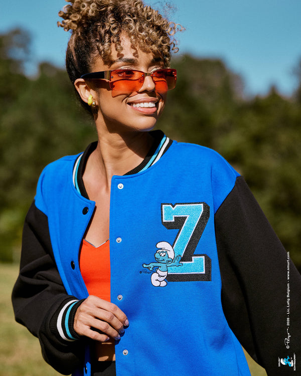 Close-up of a female model in the blue and black varsity jacket worn open over a red bra, showing the aqua blue chenille ‘Z’ with a Smurf leaning against it on the left chest, with blurred trees in the background.