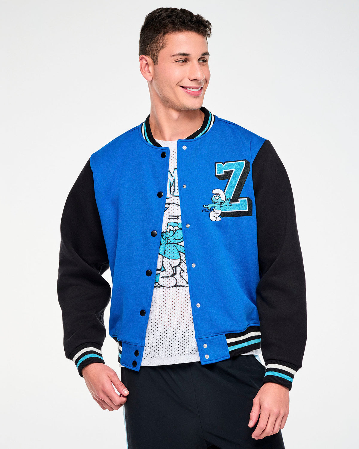 Male model in a varsity jacket with a blue fleece body, black sleeves, and black, white, and light blue striped ribbed collar, cuffs, and hem, worn open over a white mesh Smurfs graphic tee. A light blue chenille varsity 'Z' on the left chest has an embroidered Smurf leaning on it and pointing. Paired with black pants.