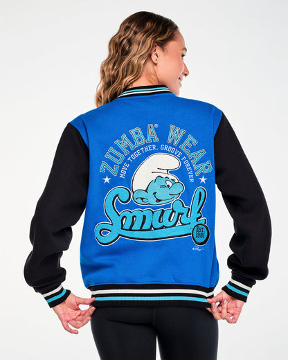 Back view of a female model in the blue and black varsity jacket, with black, white, and light blue striped ribbed trim at the collar, cuffs, and waistband. The large back graphic shows arched light blue ‘ZUMBA WEAR’ text and white ‘MOVE TOGETHER, GROOVE FOREVER’ over a chenille winking Smurf and light blue script ‘Smurf’ text, with a black ‘EST. 1981’ badge below.