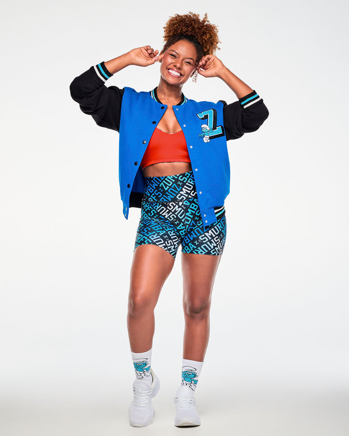 Female model with her hands at her head, wearing the blue and black varsity jacket open over a red bra, with the chenille 'Z' and Smurf on the chest, paired with blue 'ZUMBA' X 'THE SMURFS' print biker shorts, Smurf socks and white shoes.