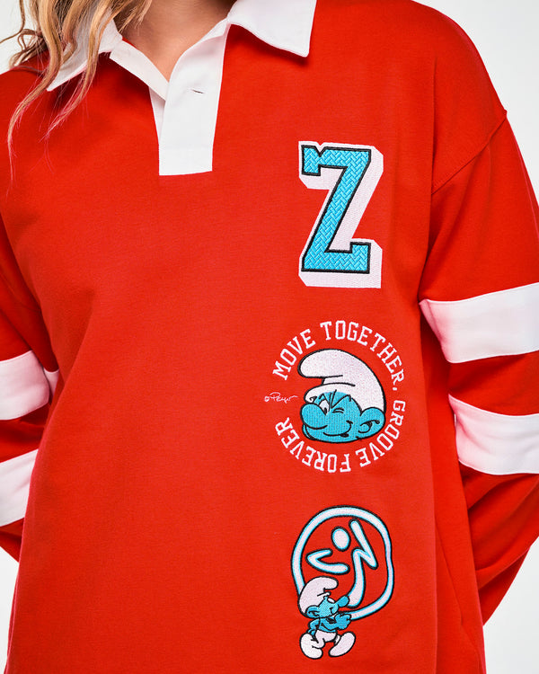 Close-up of the polo's front embroidery: a textured light blue 'Z' with white and black outlines, the winking Smurf in a circle of white text, and a Smurf walking by a blue ombre Zumbito.