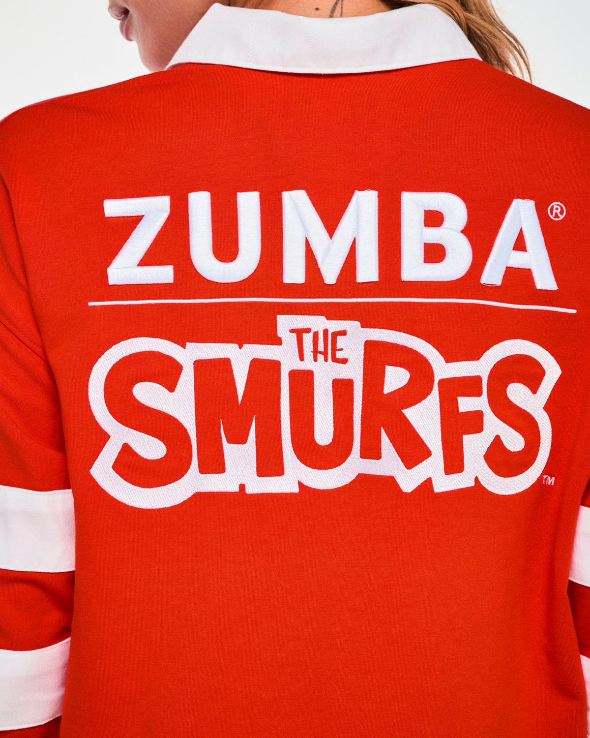 Close-up of the polo's back, showing raised white 'ZUMBA' lettering, a thin white rule, and 'THE SMURFS' logo in red letters outlined in white, just below the white collar.