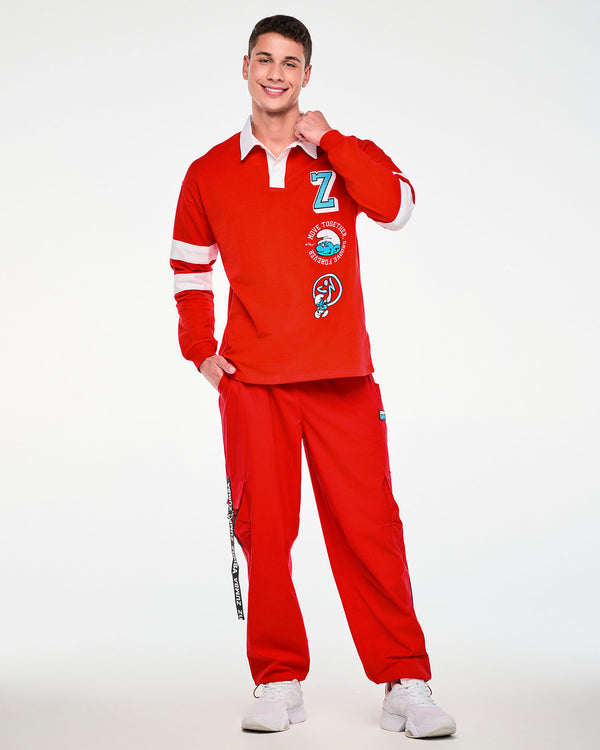 Male model holding his collar, in the red rugby polo with embroidered patches down the left front, paired with red cargo pants with black 'ZUMBA' straps.