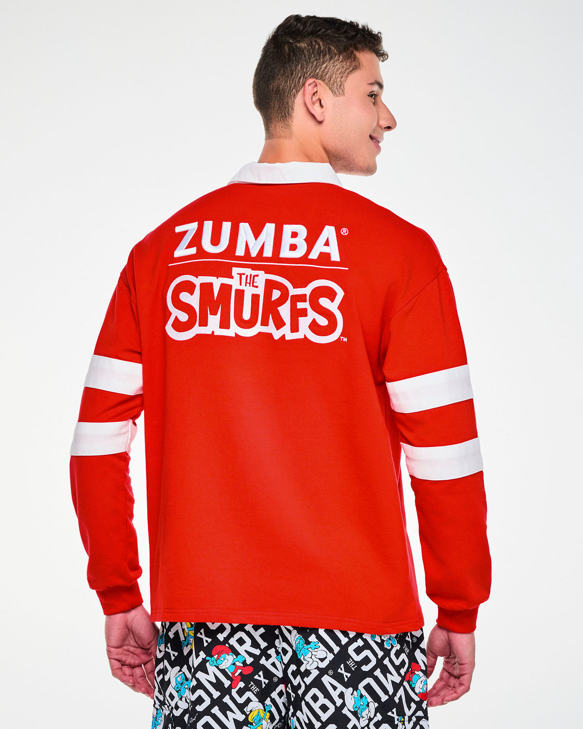 Shown from the back, a male model in the red rugby polo, with the embroidered 'ZUMBA' and 'THE SMURFS' logos across the shoulders and ribbed cuffs at the wrists.