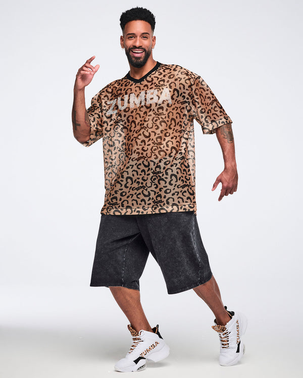 Tan leopard-print mesh top with black abstract spots, a black V-neck collar, oversized fit, and elbow-length sleeves, shown on a smiling male model wearing black acid-wash long shorts and white high-top sneakers with leopard-print details.