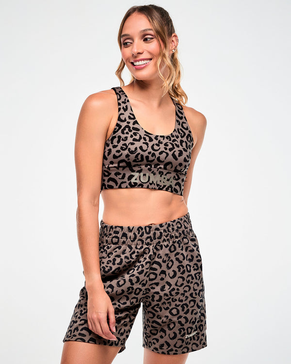 Female model in a brown racerback bra with a black leopard print, a scoop neck, and light brown 'ZUMBA' text on the underband, paired with matching leopard print shorts.