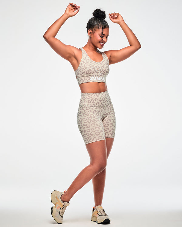 Full view of a female model with arms raised in the beige leopard print longline bra and matching high-waisted biker shorts, paired with beige shoes.