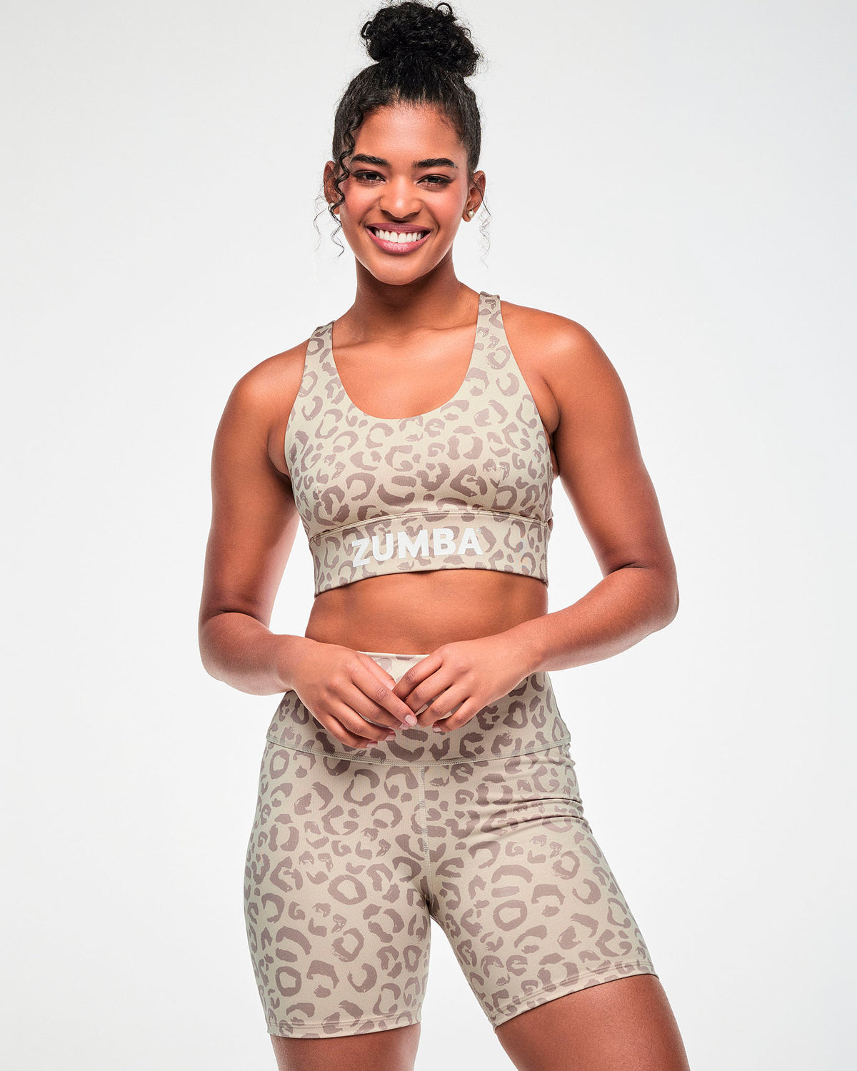 Female model in a beige longline racerback bra with a scoop neck, an all-over light brown leopard print, and white 'ZUMBA' text on the underband, paired with matching high-waisted biker shorts.