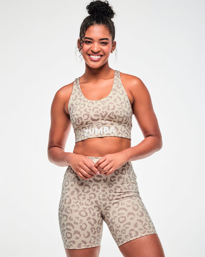 Female model in a beige longline racerback bra with a scoop neck, an all-over light brown leopard print, and white 'ZUMBA' text on the underband, paired with matching high-waisted biker shorts.