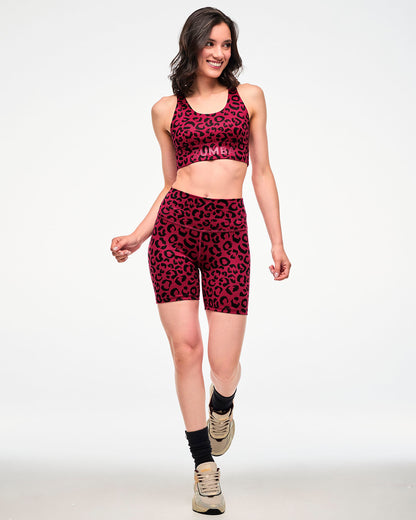 Full view of a female model walking in a dark red longline racerback bra with a black leopard print and red 'ZUMBA' text on the underband, paired with matching biker shorts, black socks, and beige shoes.