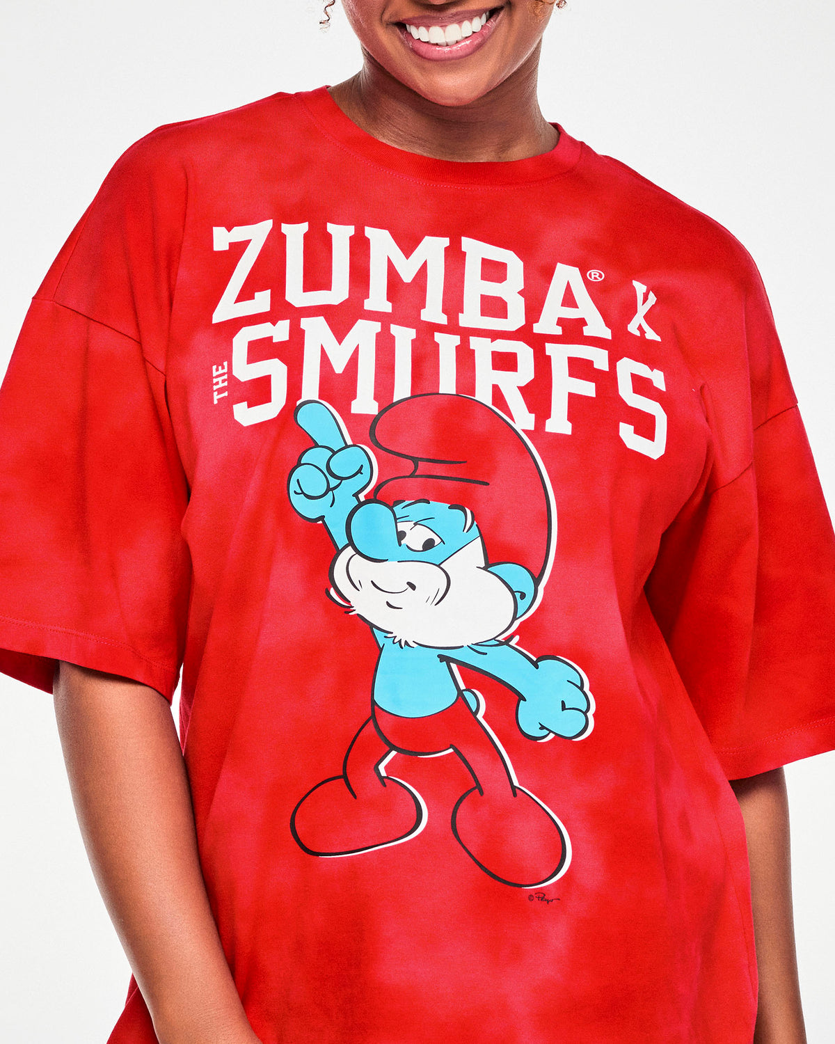 Close-up of the tee's front graphic, showing Papa Smurf in his red hat and pants with a white beard, pointing upward beneath the stacked white 'ZUMBA' X 'THE SMURFS' lettering.