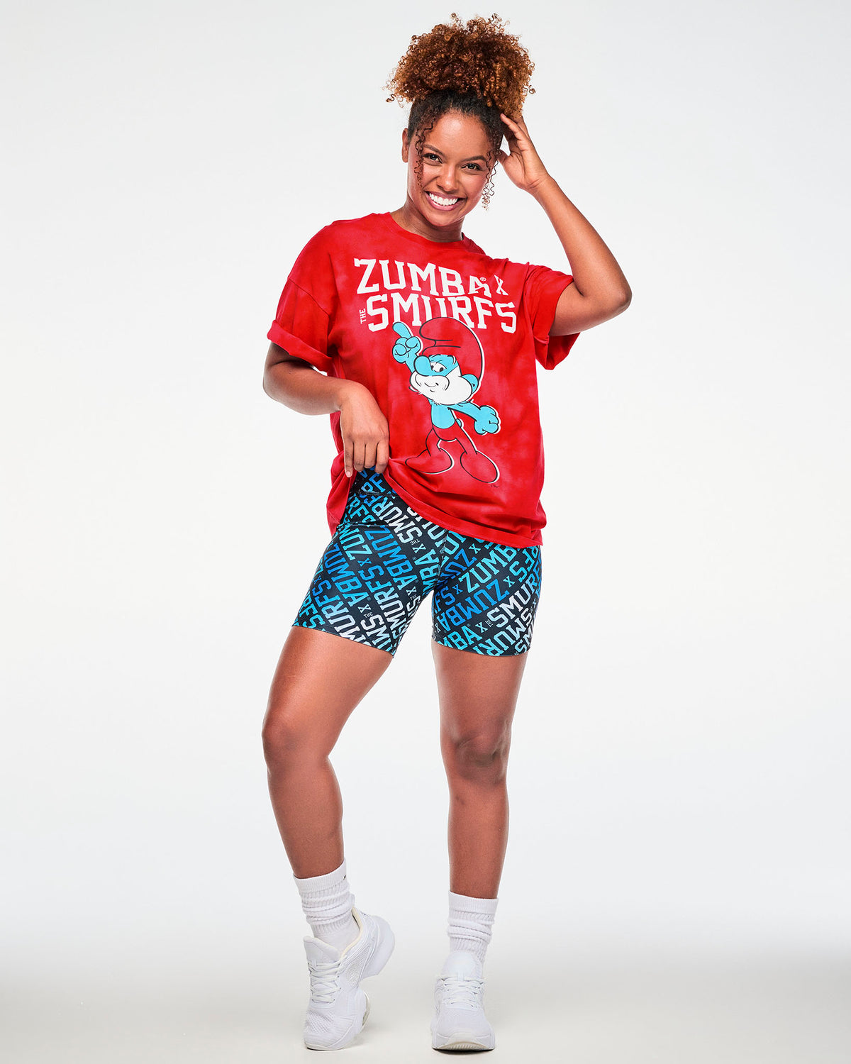 Female model with one hand in her hair, wearing the red and light cloud fabric wash tee with rolled sleeves and the white 'ZUMBA' X 'THE SMURFS' and Papa Smurf graphic, paired with blue logo print biker shorts, white socks and white shoes.