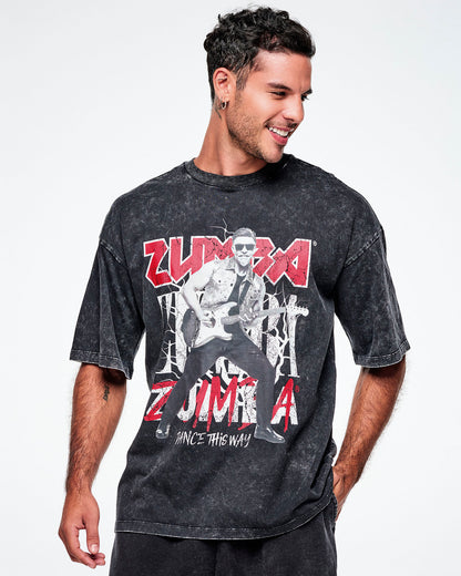A male model wearing a black acid-wash graphic tee with Beto, the creator of Zumba, smiling in sunglasses jamming on a guitar in black and white with ZUMBA in red and white text behind him and Dance this Way in smaller white text underneath, paired with red and black pants.