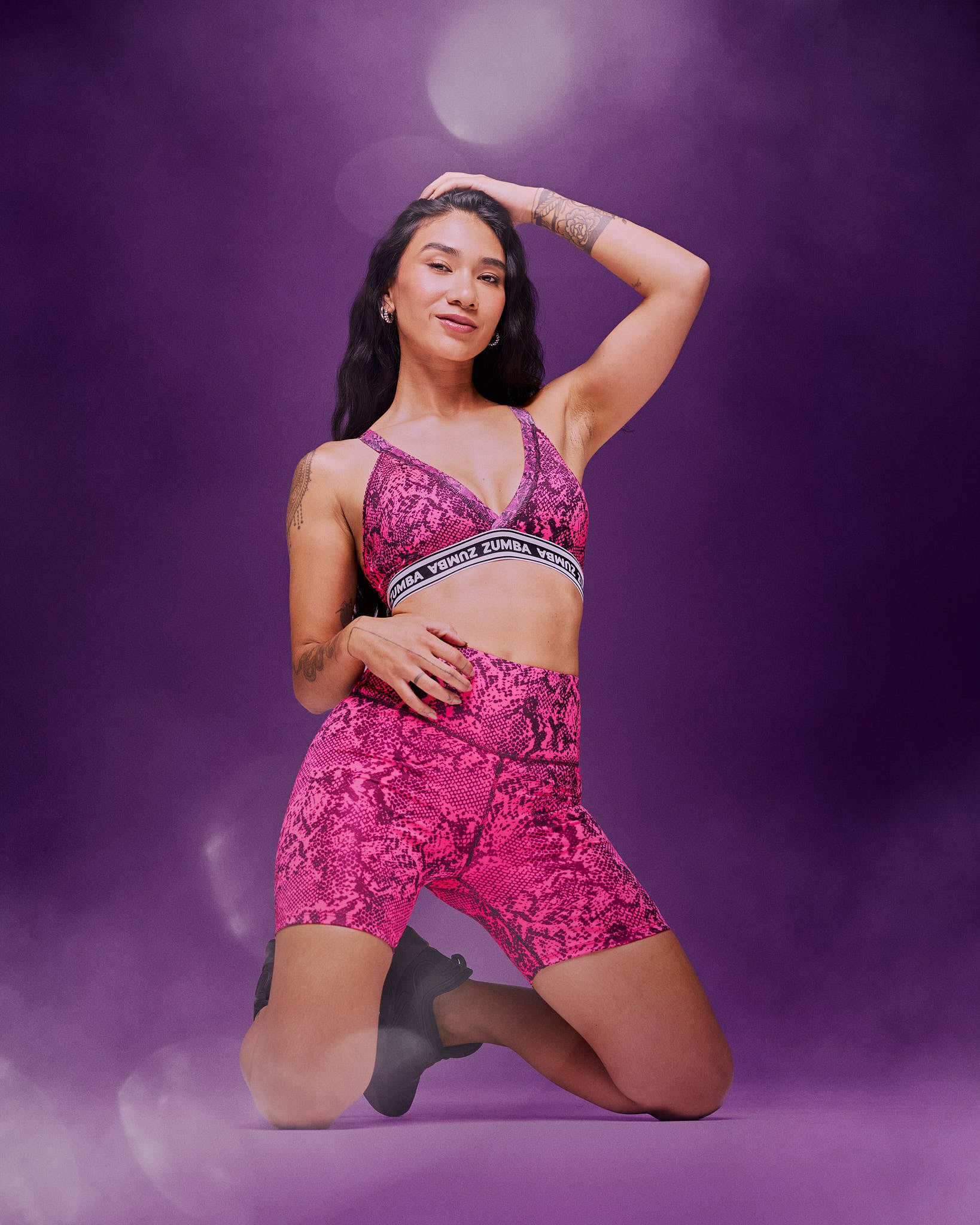 A female model wearing a bright pink patterned sports bra and matching high-waisted shorts, kneeling on the floor, against a purple studio background with smoke and light effects.
