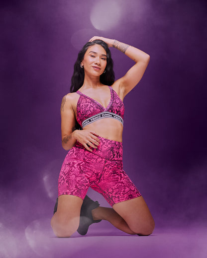 A female model wearing a bright pink patterned sports bra and matching high-waisted shorts, kneeling on the floor, against a purple studio background with smoke and light effects.