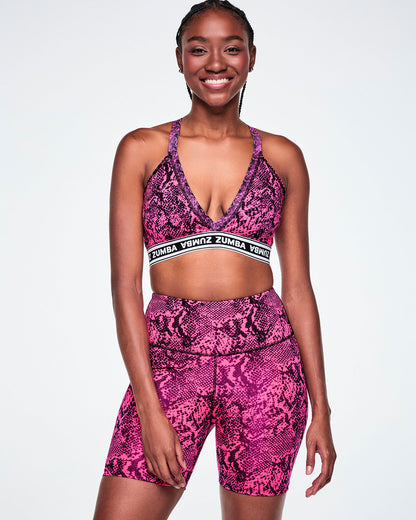 A female model wearing a pink and black snakeskin-patterned sports bra with a white and black under band printed with 'ZUMBA' in white letters and paired with matching biker shorts.