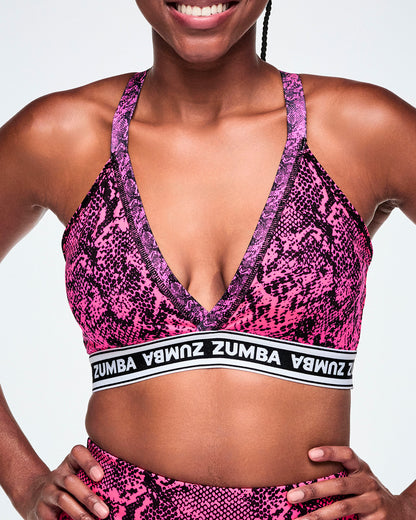 A close-up of a female model wearing a pink and black snakeskin-patterned sports bra with a white and black under band printed with 'ZUMBA' in white letters.