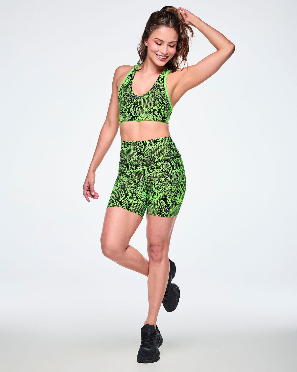 Full view of a female model wearing a matching neon green and black snakeskin print activewear set, featuring a reversible sports bra and high-waisted biker shorts, paired with black shoes.