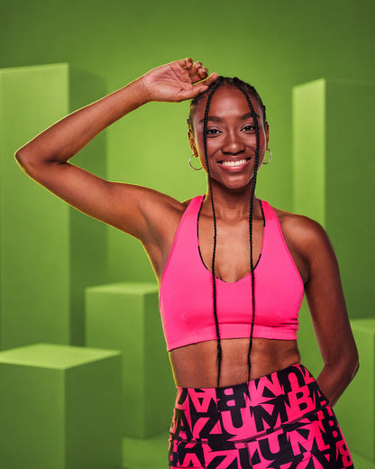 A smiling female model posing in a bright pink reversible sports bra and matching pink-and-black ZUMBA patterned athletic shorts against a green geometric studio background.