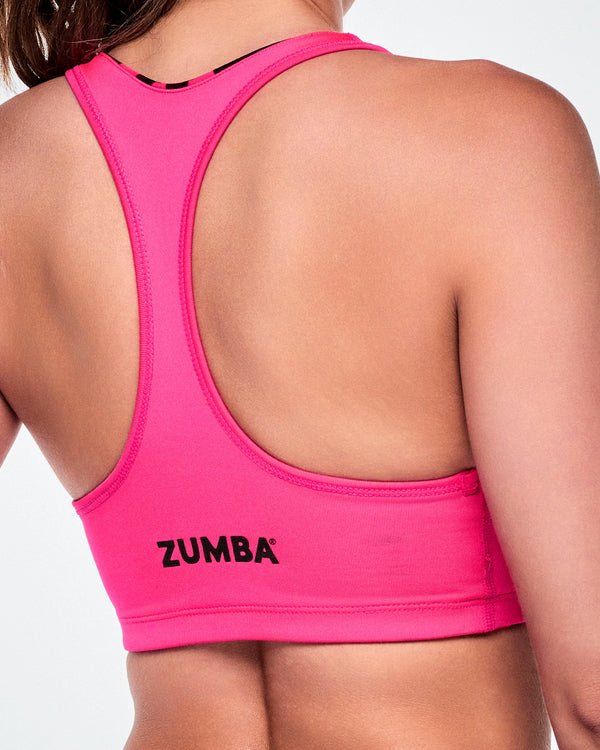 Close-up of a female model wearing a bright solid neon pink reversible sports bra with smaller black Zumba lettering towards the bottom hem.