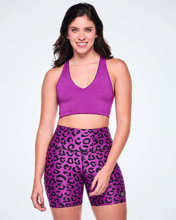 A female model wearing a solid purple reversible sports bra and matching high-waisted biker shorts with a black leopard print pattern, set against a white background.