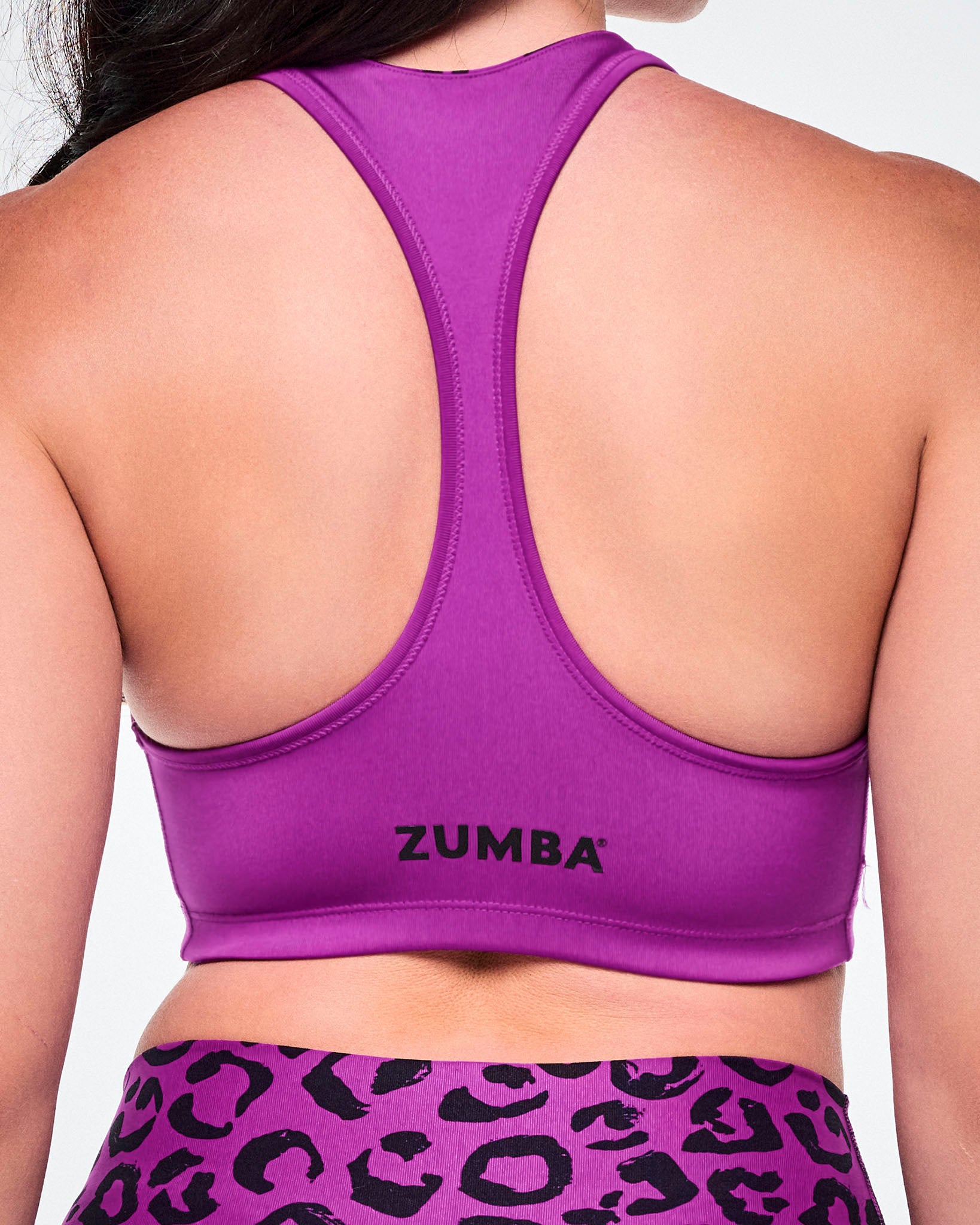 Close-up of a female model wearing a bright solid purple reversible sports bra with smaller black Zumba lettering towards the bottom hem.